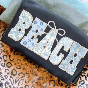 Peut inclure: T-shirt noir avec le mot "BEACH" en grandes lettres décoratives. Chaque lettre est bordée de petites perles. Les lettres sont remplies de motifs bleus et blancs sur le thème de la plage, notamment des vagues, des étoiles de mer et des feuilles tropicales.