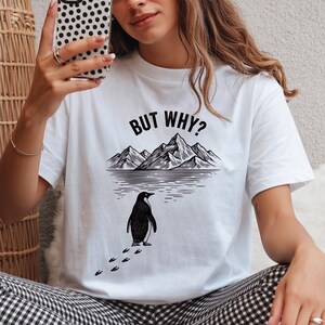 Könnte beinhalten: Weißes T-Shirt mit einem schwarz-weißen Motiv eines Pinguins, der auf eine Bergkette zugeht. Der Text "BUT WHY?" befindet sich über den Bergen. Rundhalsausschnitt.