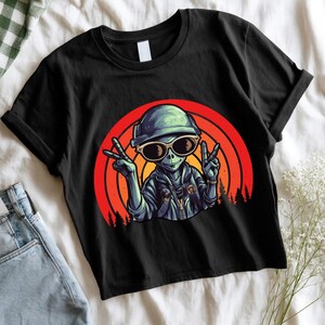 Puede incluir: Camiseta negra con un alienígena de dibujos animados con gafas de sol y un sombrero, haciendo el signo de la paz. El alienígena está sobre un fondo circular rojo y naranja con una silueta de bosque. La camiseta tiene las mangas dobladas.