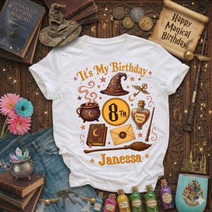 May include: White t-shirt with a colourful graphic design featuring a witch's hat, cauldron, and the text "It's My Birthday". The design includes the number "8th" and the name "Janessa". The shirt is surrounded by magical themed items.