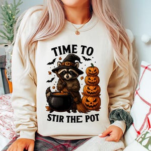 Peut inclure: Sweat-shirt beige avec un motif d'Halloween. Le dessin représente un raton laveur portant un chapeau de sorcière, remuant un chaudron, avec des citrouilles empilées et des chauves-souris. Le texte dit "TIME TO STIR THE POT".