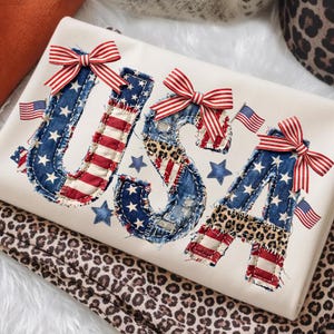 Op de afbeelding: Een crèmekleurig sweatshirt met de letters USA in een gerafeld stoffen ontwerp. De letters bevatten Amerikaanse vlagpatronen, luipaardprint accenten en rood-wit gestreepte strikken. Kleine Amerikaanse vlaggetjes en blauwe sterren zijn rond de tekst verspreid.