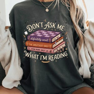 Peut inclure: Sweat-shirt gris foncé avec l'inscription "DON'T ASK ME WHAT I'M READING" et une pile de livres. Les livres sont intitulés "definitely smut", "THE SMUTTIEST SMUT" et "EVEN MORE SMUT LOL". Le motif comprend des étoiles, des lunes et des éléments décoratifs.