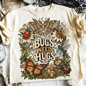 May include: A cream-colored t-shirt featuring a graphic print of various insects including a moth, ladybug, bee, praying mantis, butterfly, and beetle surrounded by flowers and mushrooms with the text BUGS NEED HUGS in the center.
