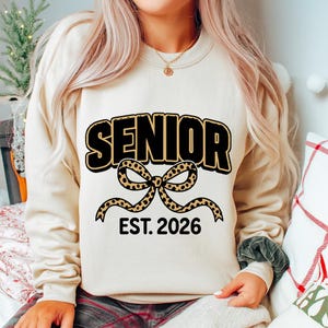 Op de afbeelding: Een beige sweatshirt met het woord "SENIOR" in zwarte blokletters met een gouden omlijning. Onder het woord "SENIOR" staat een strik met een luipaardprint. De tekst "EST. 2026" staat onder de strik.