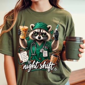 May include: An olive green t-shirt featuring a graphic of a raccoon dressed as a medical professional in green scrubs, wearing heart-shaped glasses, holding an iced coffee and a syringe, with a clipboard, stethoscope, and the text night shift.