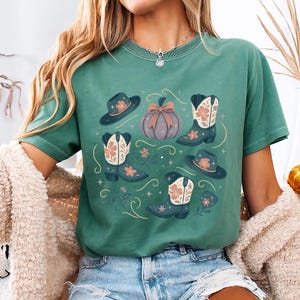 May include: Green t-shirt featuring a fall-themed design. The design includes cowboy boots, hats, a pumpkin, and floral accents in shades of blue, pink, and brown. The shirt is a comfortable, casual style.