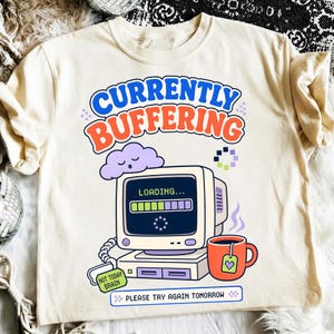Peut inclure: Un t-shirt couleur crème avec un motif d'ordinateur vintage affichant une barre de chargement, un nuage violet et une tasse orange. Le texte indique CURRENTLY BUFFERING, LOADING, NOT TODAY BRAIN et PLEASE TRY AGAIN TOMORROW en polices colorées.