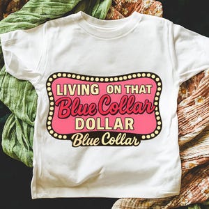 May include: White t-shirt with a pink and yellow graphic that reads "LIVING ON THAT Blue Collar DOLLAR Blue Collar". The graphic is in a retro style with a black dotted border.