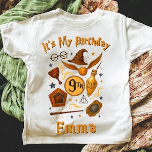 Peut inclure: T-shirt blanc avec un graphisme coloré sur le thème de la fantaisie. Le motif comprend des lunettes, un chapeau de sorcier, un vif d'or et les textes "It's My Birthday" et "Emma".