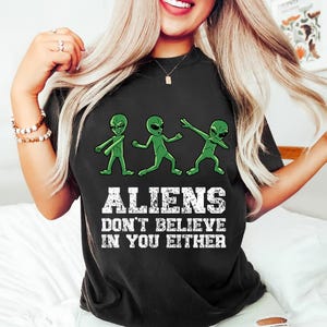 May include: Black t-shirt featuring three green cartoon aliens in various poses. The shirt has the text "ALIENS DON'T BELIEVE IN YOU EITHER" in white block letters. The shirt is a crew neck style.