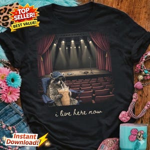 Peut inclure: T-shirt noir avec un dessin d'un raton laveur portant des lunettes de soleil et un chapeau, tenant une boisson, assis dans un fauteuil de théâtre. Le motif comprend une scène avec des rideaux rouges et le texte "i live here now."
