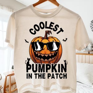 Peut inclure: T-shirt crème avec une citrouille souriante portant des lunettes de soleil. Le texte "COOLEST PUMPKIN IN THE PATCH" est en noir. Deux chats noirs et des chauves-souris sont également présents.