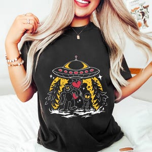 Peut inclure: T-shirt noir avec un motif fantaisiste d'un OVNI planant au-dessus d'un Bigfoot et d'un extraterrestre se tenant la main, avec un cœur entre eux. Le motif comprend un fond jaune avec des arbres et des étoiles, créant une scène amusante et ludique.