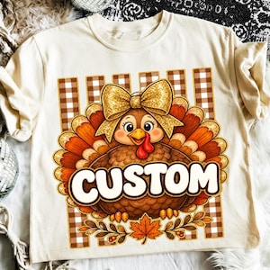 Peut inclure: Un t-shirt couleur crème avec un motif de dinde portant un nœud doré à paillettes sur la tête, devant des rayures verticales marron et blanches. Le mot CUSTOM est imprimé en grandes lettres blanches sur le corps de la dinde.