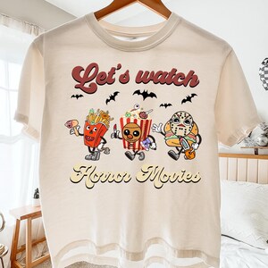 May include: Cream-colored t-shirt featuring a graphic with the text "Let's watch Horror Movies." The design includes cartoon characters of french fries, popcorn, and a masked figure, with bats above.
