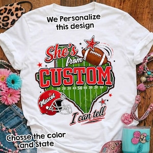 Peut inclure: Un t-shirt blanc avec un motif graphique représentant le contour d'un état en vert, un ballon de football américain marron, un casque rouge avec le mot Mascot et le texte She's from CUSTOM I can tell, agrémenté d'étoiles rouges et de lignes.