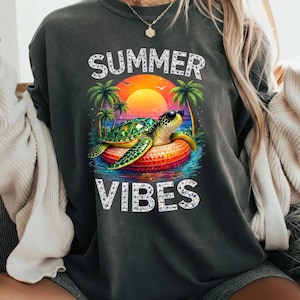 Peut inclure: Sweat-shirt gris foncé avec un graphisme coloré. Le graphisme représente une tortue portant des lunettes de soleil sur un flotteur dans l'océan, avec un coucher de soleil et des palmiers en arrière-plan. Les mots "SUMMER VIBES" sont imprimés au-dessus et en dessous de l'image.