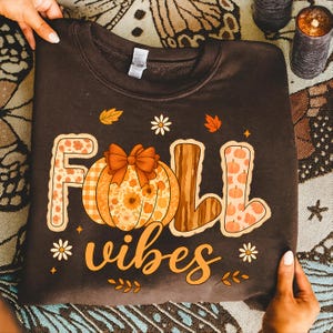 Peut inclure: Un sweat-shirt marron foncé avec les mots "FALL vibes" en écriture orange. Le mot "FALL" est décoré d'une citrouille et de motifs sur le thème de l'automne. Des fleurs blanches et des feuilles ornent le design.