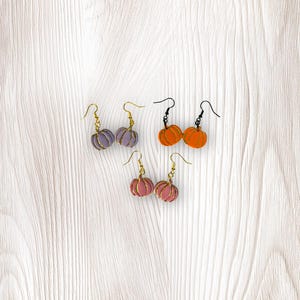 Könnte beinhalten: Vier Paar kürbisförmige Ohrringe in den Farben Lavendel, Orange und Rosa. Jeder Kürbis hat eine goldene oder schwarze Drahtkontur und einen passenden Ohrhaken. Die Ohrringe werden auf einer hellen Holzmaserung präsentiert.