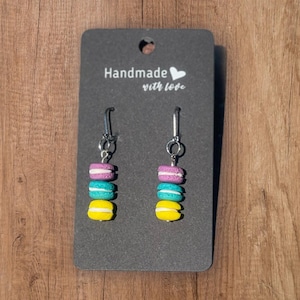 Könnte beinhalten: Ein Paar handgefertigte Ohrringe mit bunten Macaron-Anhängern in Gelb, Türkis und Lila. Die Ohrringe hängen an silberfarbenen Haken. Die Ohrringe werden auf einer grauen Karte mit dem Text "Handmade with love" präsentiert.
