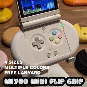 May include: A handheld gaming device in off-white with a screen displaying a colorful video game. The device has a D-pad, colored action buttons, and text that reads "4 SIZES, MULTIPLE COLORS, FREE LANYARD, MIYOO MINI FLIP GRIP".