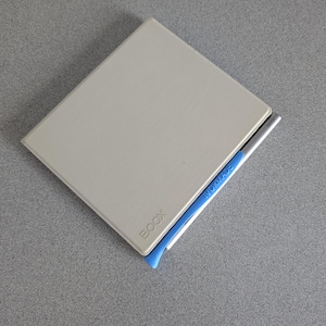 May include: A light gray BOOX e-reader with a blue stylus holder and a silver stylus. The e-reader is rectangular and has rounded corners. The stylus is silver with the word "WORMADE" printed on it.