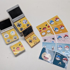 Könnte beinhalten: Gelbe und graue Handheld-Spielkonsolen mit Pikachu-Designs werden zusammen mit Stickerbögen mit verschiedenen Pokémon-Charakteren und Poké-Bällen gezeigt. Die Konsolen sind geöffnet und geschlossen und zeigen ihre Bildschirme und Tastenanordnungen. Die Aufkleber zeigen verschiedene Pokémon.