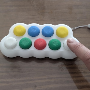 May include: A white, cloud-shaped fidget toy with eight colorful, raised buttons in yellow, blue, green, and red. A finger presses one of the white buttons. A white lanyard is attached to the toy.