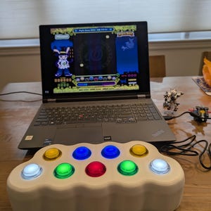 May include: A white, cloud-shaped gaming controller with eight multi-colored, illuminated buttons. A laptop displaying a video game is behind the controller. The buttons are yellow, blue, green, and red. A small toy is to the right of the laptop.