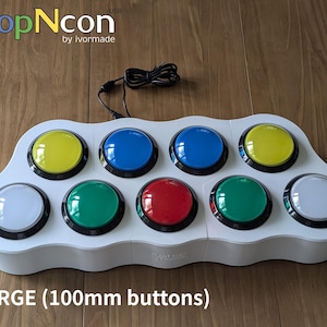 May include: A white electronic device with ten large, colorful buttons. The buttons are yellow, blue, green, red, and white. The device has the text "PopNcon" and "LARGE (100mm buttons)". A black cable extends from the device.