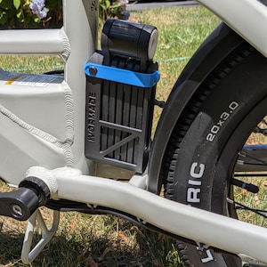 May include: A black folding bicycle lock with a blue strap mounted to the frame of a light gray electric bicycle. The lock features the text IVORMADE on its side. The bicycle tire is marked with the text 20x3.0, indicating its size.