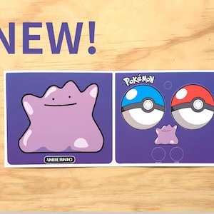 May include: A sticker sheet featuring Pokémon characters. The sheet includes a purple Ditto, two Poké Balls, and the word "NEW" in purple. The brand name "ANBERNIC" is also visible.