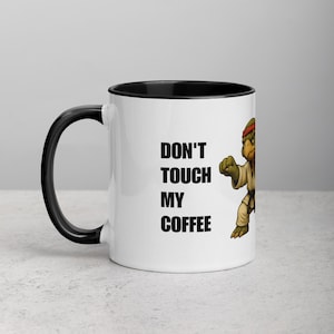 May include: White ceramic coffee mug with a black interior and handle. The mug features the text "DON'T TOUCH MY COFFEE" in black, along with an illustration of a turtle in a karate uniform.