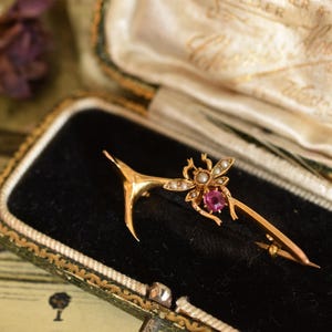 May include: A gold brooch in a vintage jewellery box. The brooch features a stylized bee design with small pearls and a central pink gemstone. The box is lined with black velvet and has a green and gold trim.