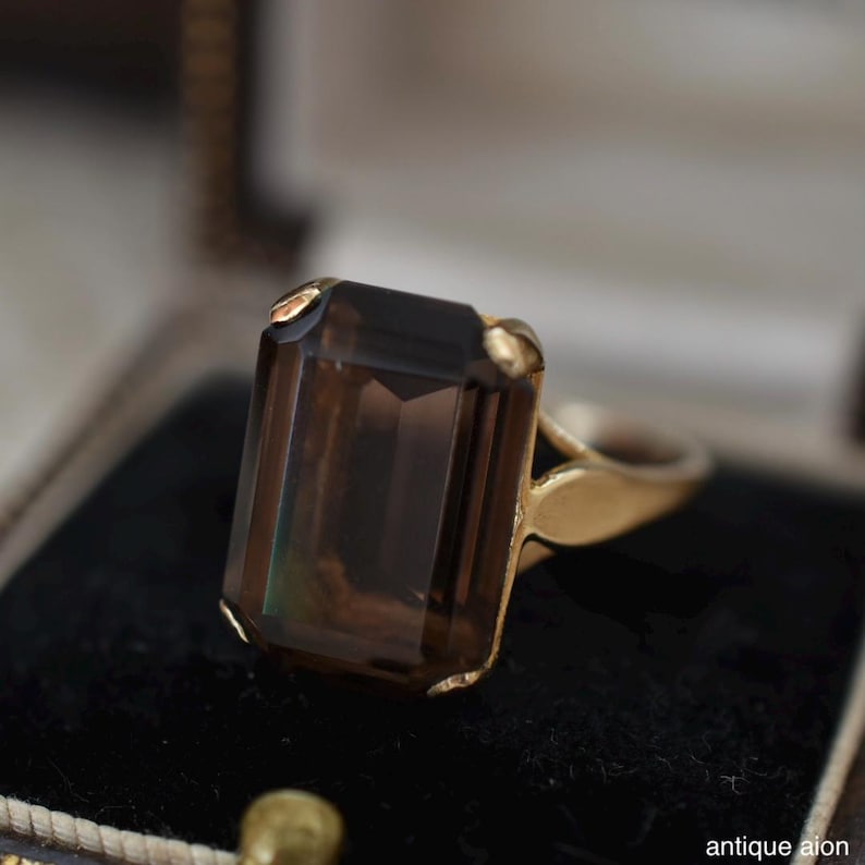 May include: A gold ring featuring a large, rectangular, smoky brown gemstone. The ring's setting is a warm gold color, and the gemstone has a faceted cut. The ring is displayed on a black velvet surface.