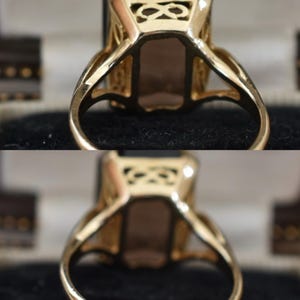 May include: A gold ring with a large, rectangular, dark gemstone. The ring has an ornate, openwork design on the shoulders and around the gemstone setting. The band is smooth and polished. The ring is set against a dark background.