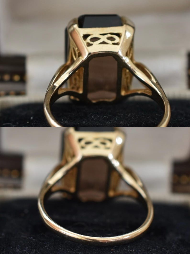 May include: A gold ring with a large, rectangular, dark gemstone. The ring has an ornate, openwork design on the shoulders and around the gemstone setting. The band is smooth and polished. The ring is set against a dark background.