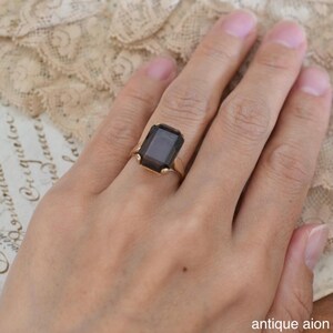 May include: A gold-toned ring featuring a large, rectangular, smoky-gray gemstone. The ring has a classic design with a simple band and prongs holding the stone. The image is set against a soft, neutral background. The text "antique aion" is in the bottom right corner.