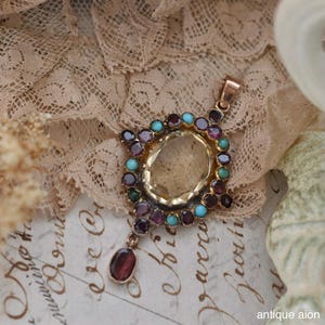 Peut inclure: Pendentif ancien en métal doré avec une grande pierre ovale jaune clair au centre, entourée de petites pierres rondes violettes, turquoise et vertes. Une seule pierre rouge ovale est suspendue en dessous. Le pendentif repose sur de la dentelle vintage.