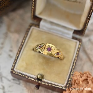 May include: A gold ring with a central square setting featuring a ruby and star design, with smaller ruby and diamond accents. The ring is presented in a vintage jewellery box with a velvet interior, highlighting its antique style.