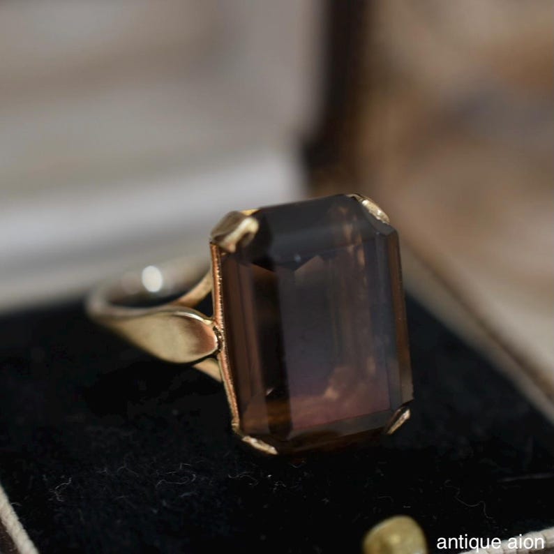 May include: A gold ring featuring a large, rectangular, smoky brown gemstone. The ring's band is a light gold color, and the setting around the gemstone is a darker gold. The ring is displayed on a black surface. The text "antique aion" is visible.