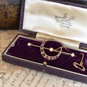 Puede incluir: Broche dorado con forma de media luna, con piedras rojas y una perla central, expuesto en una caja forrada de terciopelo morado. El broche tiene un alfiler recto con detalles de perlas y una cadena de imperdible.