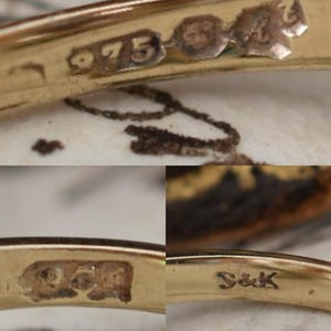 May include: Close-up of a gold ring with several hallmarks. The ring is stamped with "975" and other markings. The ring's surface is reflective, and the details are visible against a blurred background.