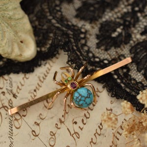 Puede incluir: Horquilla para el pelo con forma de araña dorada con cuerpo de piedra turquesa, acentuada con rubí y esmeralda. La araña está montada sobre una fina barra rectangular dorada. El fondo incluye encaje y papel vintage.