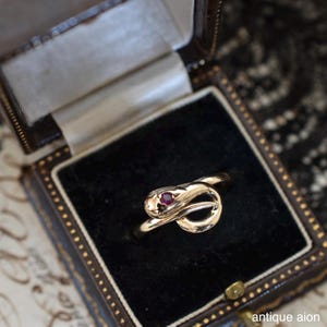 May include: A gold snake ring with a ruby eye, displayed in a vintage jewellery box. The ring has a coiled snake design. The box is lined with black velvet.