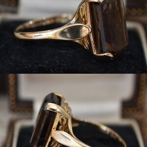 May include: A gold ring featuring a large, rectangular, smoky brown gemstone. The ring has an ornate, detailed setting and a polished gold band. The gemstone is faceted, catching the light and displaying a rich, dark color. The ring is displayed in a jewelry box.