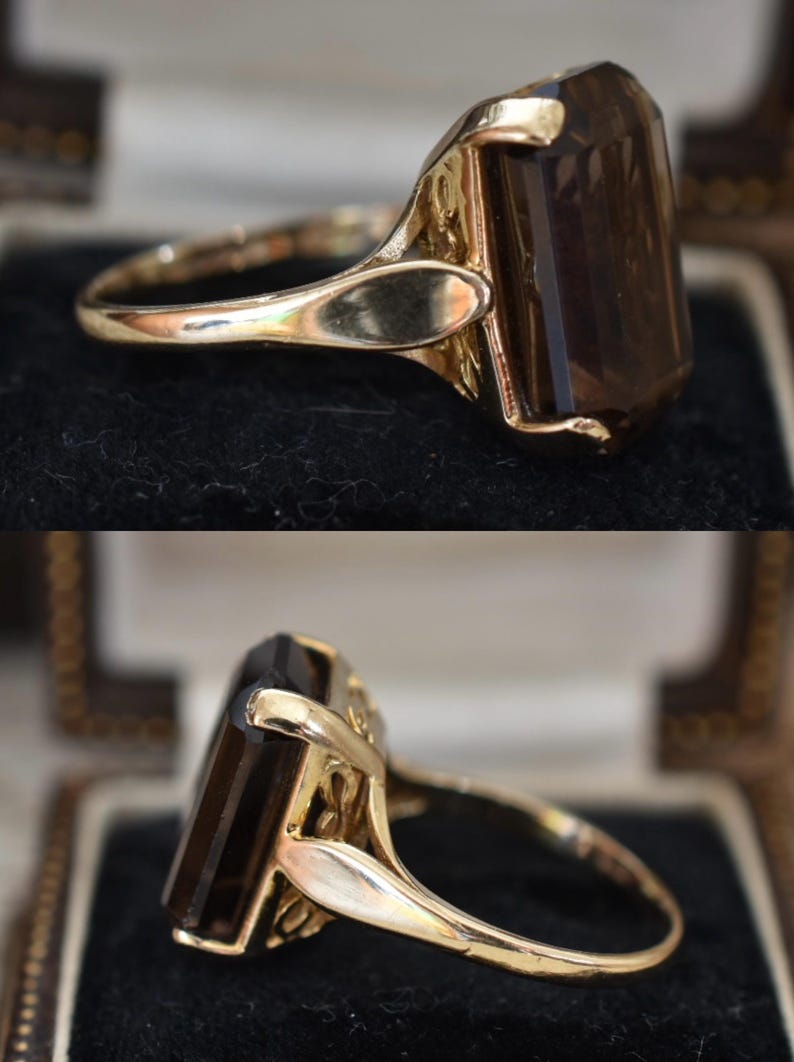 May include: A gold ring featuring a large, rectangular, smoky brown gemstone. The ring has an ornate, detailed setting and a polished gold band. The gemstone is faceted, catching the light and displaying a rich, dark color. The ring is displayed in a jewelry box.