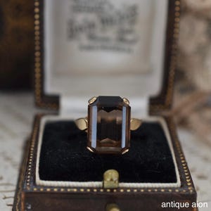 May include: A vintage gold ring with a large, rectangular, smoky brown gemstone. The ring is set in a classic style and displayed in a brown jewelry box with a black velvet interior. The text "antique aion" is visible.