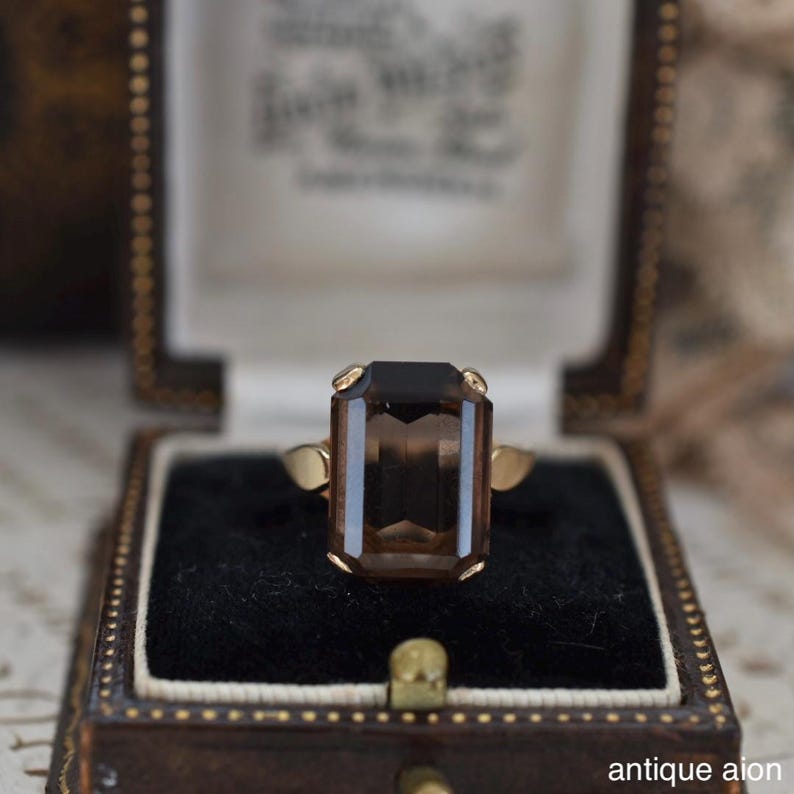 May include: A vintage gold ring with a large, rectangular, smoky brown gemstone. The ring is set in a classic style and displayed in a brown jewelry box with a black velvet interior. The text "antique aion" is visible.